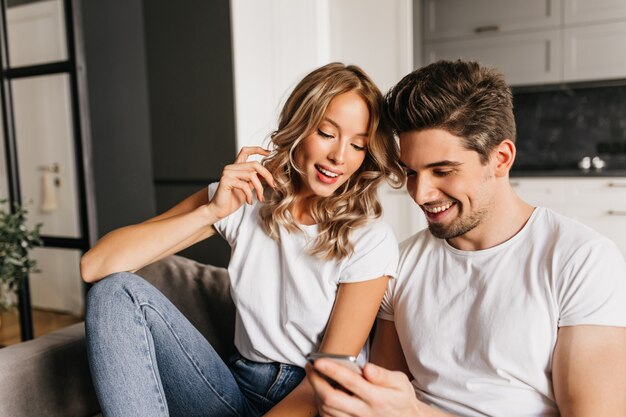 Gelukkige paar met slimme telefoon kijken op het scherm en glimlachen. Huisportret van twee jonge mensen die van dag samen genieten en goed nieuws lezen.