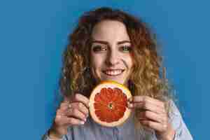 Gratis foto gelukkige positieve jonge 25-jarige vrouw met golvend kapsel met plakje verse grapefruit, handen reiken alsof ze je sappig vitamine fruit aanbieden. gezonde levensstijl en fruitarisme concept