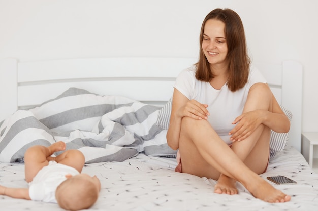 Gelukkige positieve vrouw met een wit t-shirt en korte broek zittend op bed bedekt wit laken met haar dochtertje, donkerharige vrouw die met een glimlach naar haar lieftallige kind kijkt.