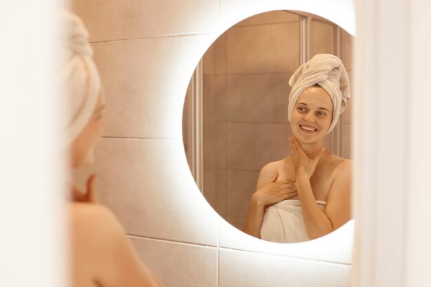 Gelukkige positieve vrouw met frisse huid die na het douchen in de spiegel kijkt, staand in een witte handdoek gewikkeld, poserend in de badkamer terwijl ze ochtendprocedures uitvoert.