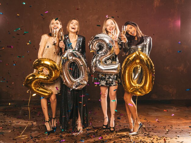 Gelukkige prachtige meisjes in stijlvolle sexy feestjurken met gouden en zilveren 2020-ballonnen