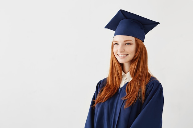 Gelukkige roodharige gediplomeerde vrouw die over witte oppervlakte glimlacht