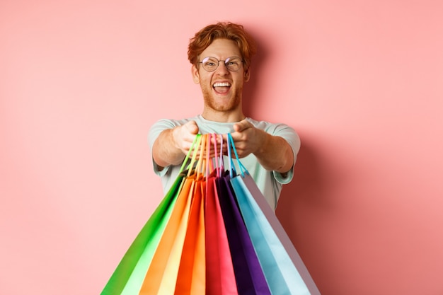 Gelukkige roodharige man strekt zijn handen uit met boodschappentassen, geeft je cadeaus, staande over roze achtergrond.