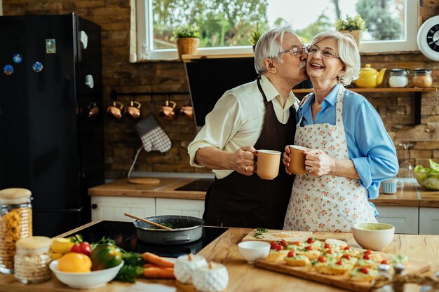 Gelukkige senior man kust zijn vrouw terwijl hij koffie drinkt en eten klaarmaakt met haar in de keuken
