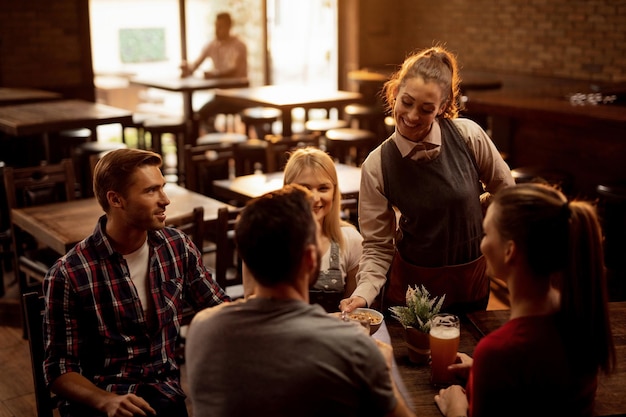 Gelukkige serveerster die pinda's serveert aan een groep jonge mensen die bier drinken in een pub