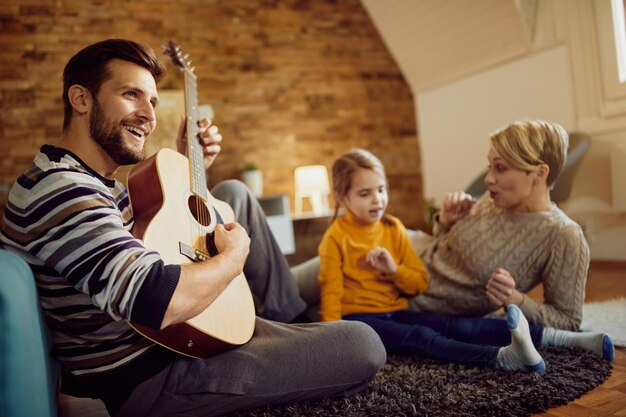 Gelukkige vader die akoestische gitaar speelt terwijl hij thuis ontspant met zijn gezin