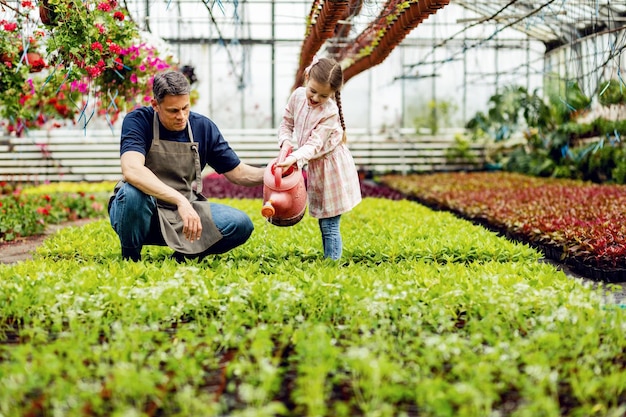 Gelukkige vader en dochter die plezier hebben tijdens het gebruik van gieter en voedende bloemen op plantenkwekerij