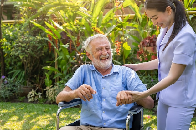 Gelukkige verpleegster die lachende oudere man hand op rolstoel vasthoudt in tuin bij verpleeghuis