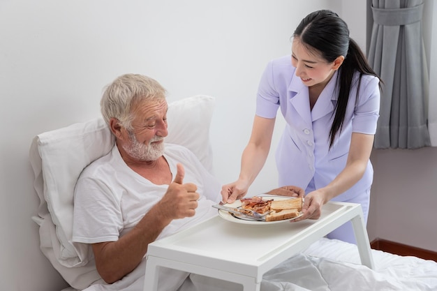 Gelukkige verpleegster serveert bejaarde senior man ontbijt op bed in slaapkamer bij verpleeghuis duim omhoog