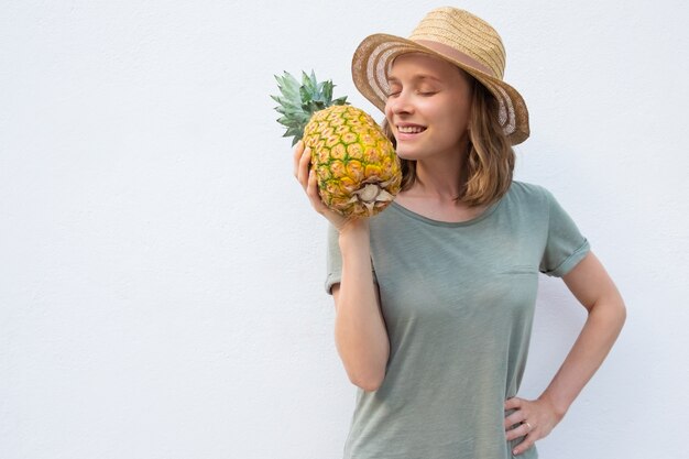 Gelukkige vreedzame vrouw die in de zomerhoed gehele ananas ruikt