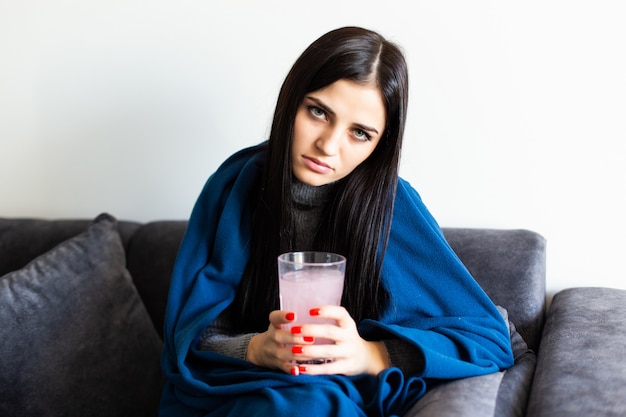 Gelukkige vrouw die een witte ronde pil en een glas water houdt bekijkend camerazitting op een bank