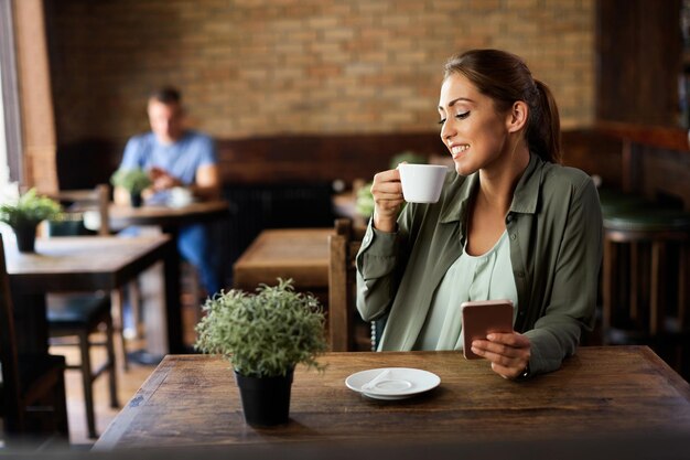 Gelukkige vrouw die geniet van een kopje koffie tijdens het gebruik van een smartphone in een café