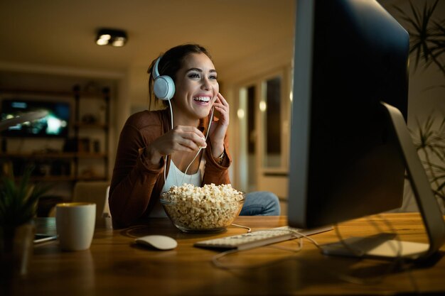 Gelukkige vrouw die popcorn eet en film kijkt op desktop-pc terwijl ze 's avonds geniet in haar appartement
