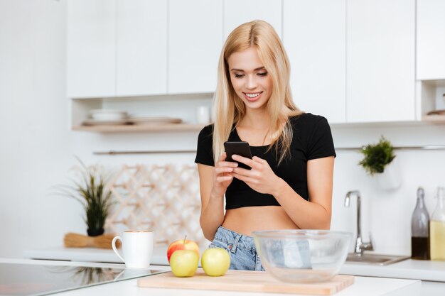 Gelukkige vrouw die telefoon op keuken met behulp van