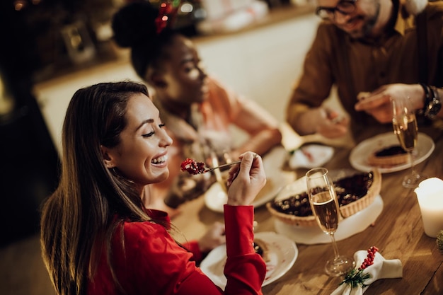 Gelukkige vrouw die vrolijke taart eet terwijl ze thuis een kerstdiner heeft met vrienden