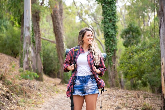 Gelukkige vrouw die zich op bosweg bevindt, glimlacht en wegkijkt. Langharige vrouw met rugzakken en wandelen op de natuur. Toerisme, avontuur en zomervakantie concept