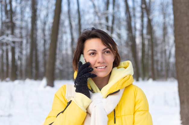 Gelukkige vrouw in een goed humeur wandelingen door het besneeuwde winterbos en vrolijk chatten aan de telefoon, genieten van tijd buiten in het park