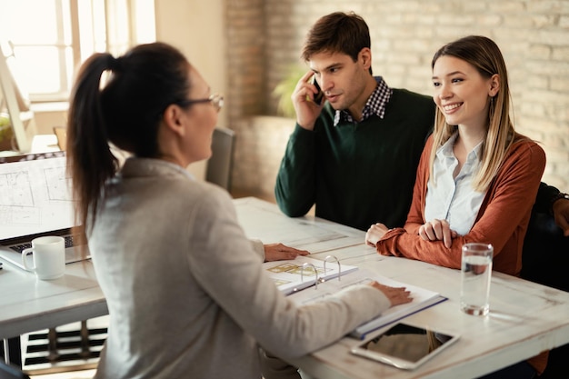 Gelukkige vrouw in gesprek met financieel adviseur terwijl haar man aan het telefoneren is tijdens een vergadering op kantoor