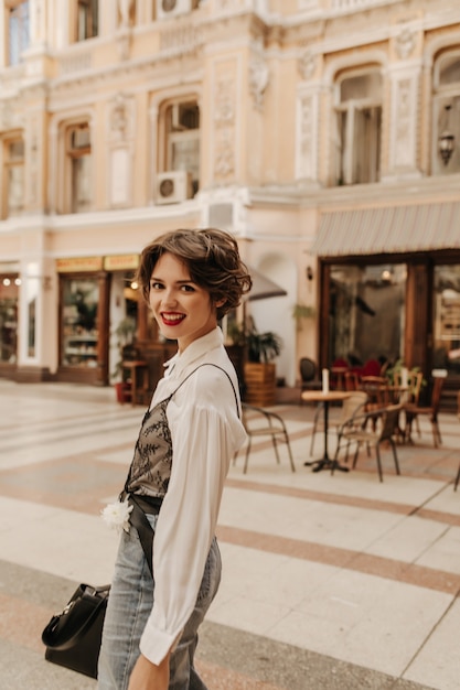 Gelukkige vrouw in licht overhemd en spijkerbroek in de stad. Moderne vrouw met kort haar en heldere lippen lachend op straat.