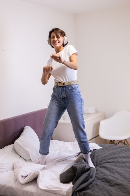 Gelukkige vrouw in vrijetijdskleding thuis in slaapkamer luistert naar muziek in hoofdtelefoons, dansen en springen