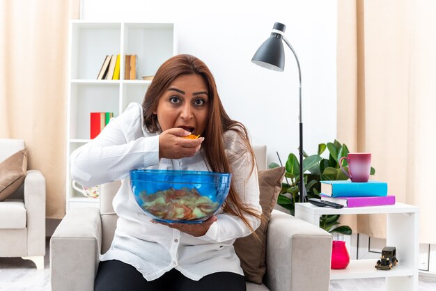 Gelukkige vrouw in wit overhemd en zwarte broek ontspannen zittend op de stoel met kom chips eten chips in lichte woonkamer living