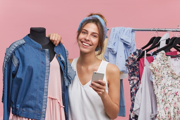 Gelukkige vrouw met een aantrekkelijk uiterlijk, staande in de buurt van etalagepop, met een vreugdevolle uitdrukking terwijl ze blij is om een nieuwe outfit te kopen, berichten typt naar haar beste vriendin, het laatste nieuws met elkaar deelt