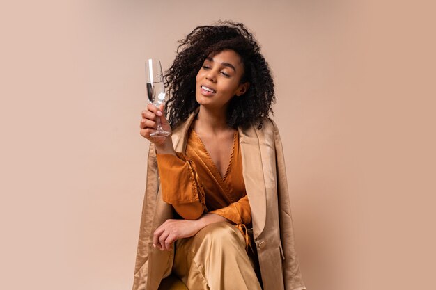 Gelukkige vrouw met gebruinde huid met perfect krullende haren met glas wijn, ielegant oranje blouse en zijden broek zittend op vintage stoel beige muur.