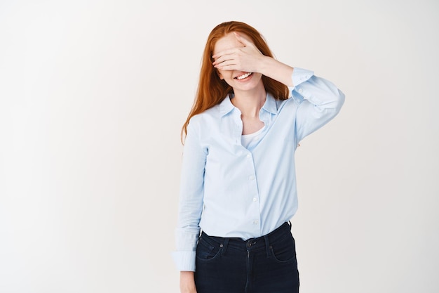 Gelukkige vrouw met gemberhaar die de ogen bedekt met de hand en zorgeloos glimlacht, wachtend op verrassing, staande over een witte muur