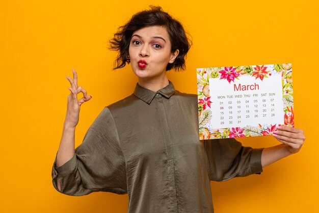 Gelukkige vrouw met kort haar die een papieren kalender van de maand maart vasthoudt en op zoek is naar een goed teken dat de internationale vrouwendag op 8 maart viert