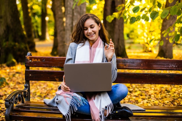 Gratis foto gelukkige vrouw praten online in een videoconferentie met een laptop zittend op een bankje in het najaar park