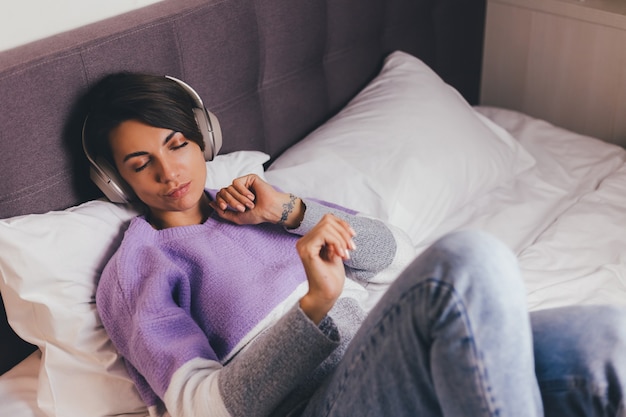 Gelukkige vrouw thuis op comfortabel bed, het dragen van warme kleren pullover, muziek luisteren