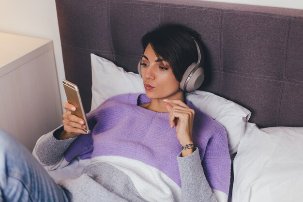 Gelukkige vrouw thuis op comfortabel bed, het dragen van warme kleren pullover, muziek luisteren