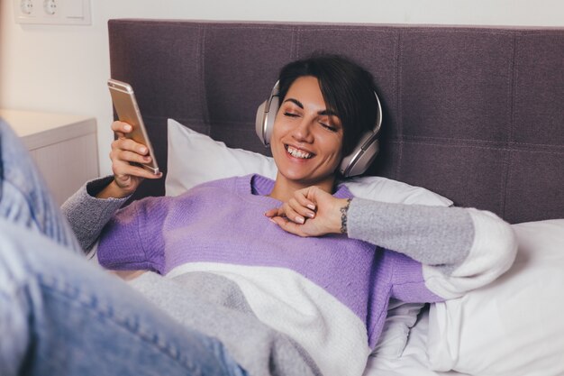 Gelukkige vrouw thuis op comfortabel bed, het dragen van warme kleren pullover, muziek luisteren