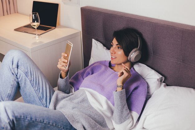 Gelukkige vrouw thuis op comfortabel bed, het dragen van warme kleren pullover, muziek luisteren