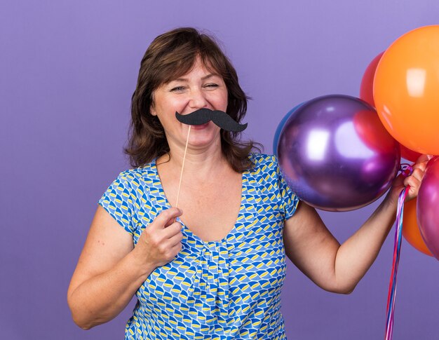 Gelukkige vrouw van middelbare leeftijd met een stel kleurrijke ballonnen en grappige snor op stok die plezier heeft