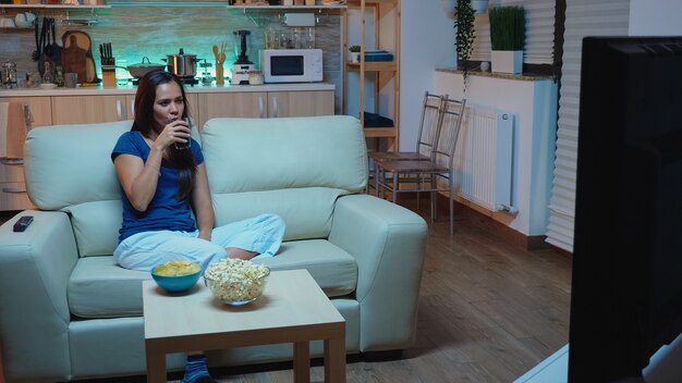 Gelukkige vrouw zittend op de bank film kijken op televisie thuis. Opgewonden, geamuseerde, eenzame dame in pijama's genietend van de avond zittend op een comfortabele bank voor de televisie popcorn etend.