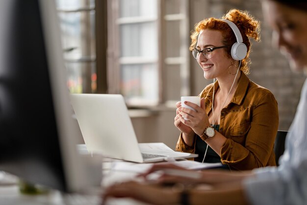 Gelukkige vrouwelijke ondernemer die een koptelefoon draagt tijdens het surfen op het net op een laptop en koffie drinkt op kantoor