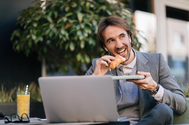 Gelukkige zakenman die croissant eet terwijl hij naar muziek luistert op oortelefoons in het buitencafé