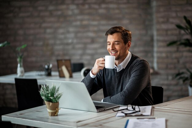 Gelukkige zakenman die koffie drinkt en iets op internet leest terwijl hij een laptop gebruikt op zijn bureau