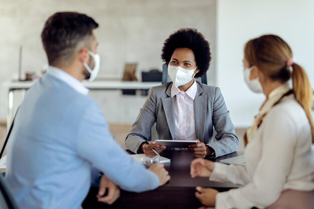 Gelukkige zwarte bankmanager die gezichtsmaskers draagt terwijl hij overleg heeft met een stel op kantoor