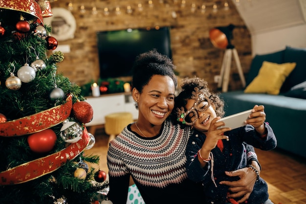 Gelukkige zwarte moeder en dochter die plezier hebben tijdens het nemen van selfie bij de kerstboom