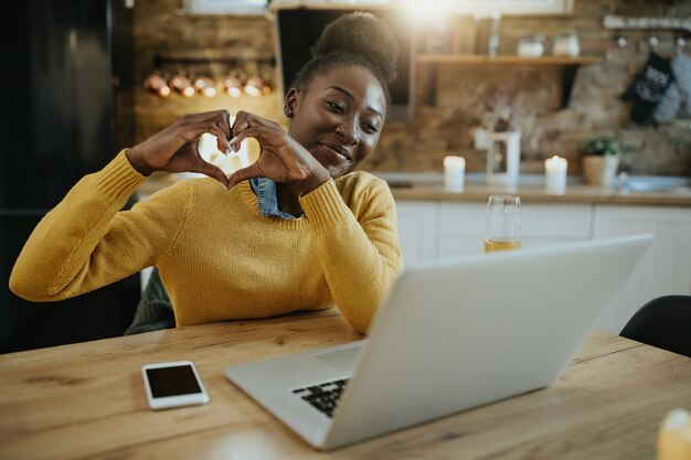 Gelukkige zwarte vrouw die hartvorm toont aan iemand tijdens videogesprek via laptop thuis