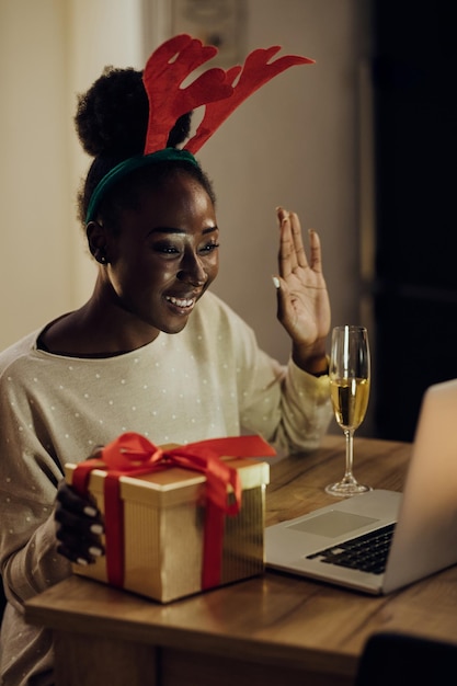 Gelukkige zwarte vrouw die laptop gebruikt en iemand begroet tijdens videogesprek op kerstavond thuis