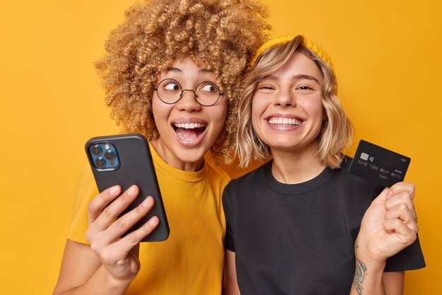 Gemakkelijke betalingen Gelukkige jonge vrouwen maken internet winkelen gebruik van mobiele telefoon en bankkaart kopen dingen online glimlach breed gekleed in casual t-shirt geïsoleerd over levendige gele achtergrond