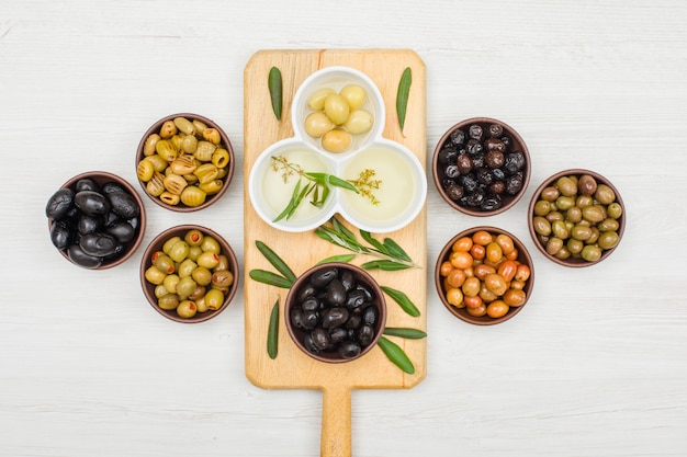 Gemarineerde olijven en olijfolie in een kommen en snijplank met olijfbladeren bovenaanzicht op wit hout