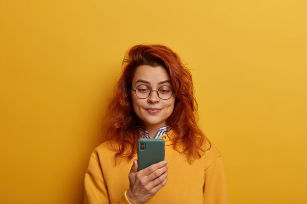 Gember jonge vrouw houdt mobiele telefoon, bericht leest, draagt ronde bril en gele trui
