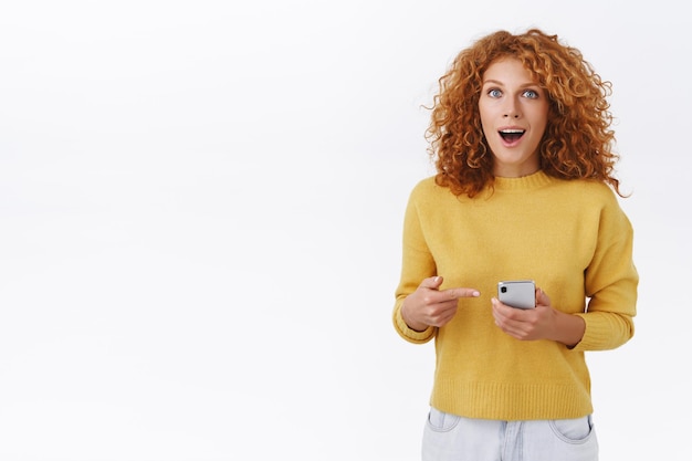 Gember meisje met krullend natuurlijk kapsel, smartphone vasthoudend, mobiele telefoon wijzend en starend naar voren onder de indruk