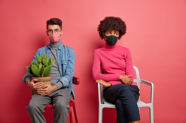 Gratis foto gemengd ras jonge vrouw en man dragen beschermende maskers hebben een slecht humeur naast elkaar zitten houdt cactus