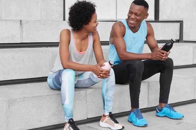 Gemotiveerde vrolijke donkere vrouw en man zitten op trappen, kijken elkaar positief aan, houden flessen water vast, dragen sportkleding en gympen, hebben discussie over sportcompetities.