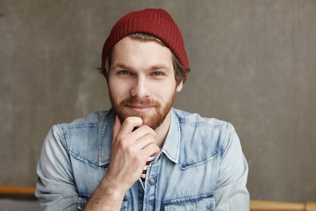 Genieten van een mooie dag. portret van vrolijke aantrekkelijke bebaarde hipster in goed humeur met rust binnenshuis, zittend tegen grijze betonnen muur in moderne café, ontspannen en zorgeloos gevoel